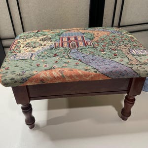 Puede incluir: Un reposapiés rectangular con cuatro patas de madera torneada de color marrón oscuro y una parte superior de tela tipo tapiz. La tapicería presenta una escena paisajística tejida con una casa, un camino azul sinuoso, árboles y motivos florales.