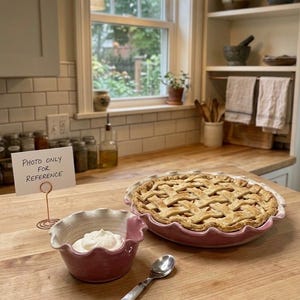 May include: A round pie with a golden-brown lattice crust in a pink fluted ceramic dish, accompanied by a small matching pink bowl containing white whipped cream and a silver spoon with a white handle, plus a sign reading Photo only for reference.