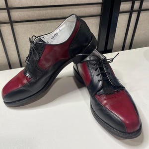 May include: A pair of two-tone black and burgundy leather lace-up shoes featuring a kiltie fringe detail over the vamp, black laces, and a low heel. The interior lining is white with visible text reading 46479D printed on the inner side.