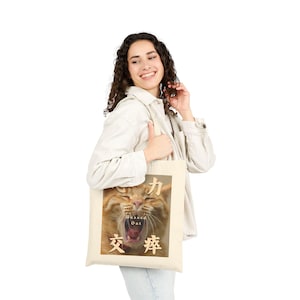 Op de afbeelding: Beige draagtas met een close-up van een geeuwende oranje tabbykat. De afbeelding bevat Japanse karakters en de woorden "Banned Out". De tas wordt vastgehouden door een persoon die een witte jas en een blauwe jeans draagt.