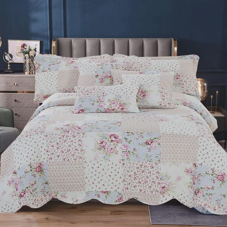 May include: A patchwork quilt and matching pillow shams featuring a floral pattern with pink roses and small blue flowers on a white background. The bedding set has a scalloped edge design and covers a bed with a gray upholstered headboard.