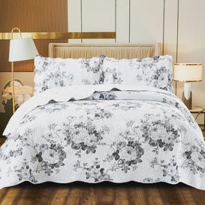 May include: A white bedspread and two matching pillow shams featuring a dark gray floral pattern. The bedding set has a quilted texture with a scalloped edge design, covering a bed frame with a headboard in a bedroom setting with two lamps.