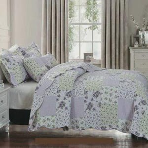 May include: A patchwork quilt and matching pillow shams featuring a floral pattern in shades of lavender, light green, and white. The bedding set displays a repeating block design with scalloped edges, covering a bed with a white fitted sheet.