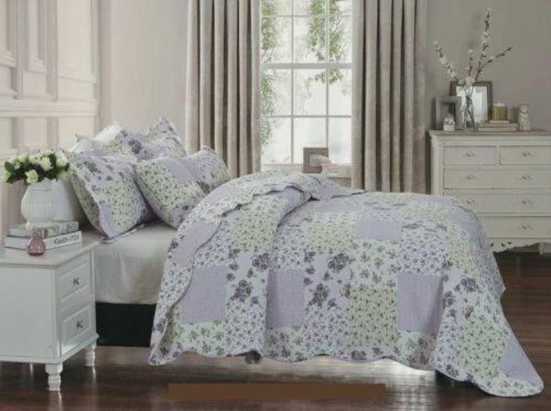 May include: A patchwork quilt and matching pillow shams featuring a floral pattern in shades of lavender, light green, and white. The bedding set displays a repeating block design with scalloped edges, covering a bed with a white fitted sheet.
