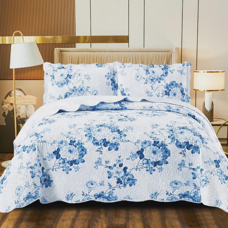 May include: A white bed quilt and two matching pillow shams featuring a blue floral pattern. The bedding set has a quilted texture with a repeating design of blue roses and leaves across the entire surface, creating a cohesive look for the bed.