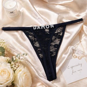 May include: Black lace thong underwear featuring a thin waistband adorned with silver-toned rhinestone letters spelling out the name DAMON. The garment is crafted from delicate floral patterned lace and includes a solid black fabric trim.