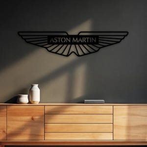 May include: A black metal wall art piece featuring the Aston Martin logo with its signature winged design and the text ASTON MARTIN centered in a rectangular frame. The piece is mounted on a dark wall above a light-colored wooden cabinet.