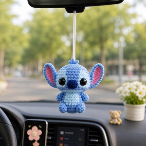 May include: A blue and light blue crochet Stitch car charm hanging from a car's rearview mirror. The charm has large ears with pink inner ears, black eyes, and a small nose. The charm is suspended by a white string.