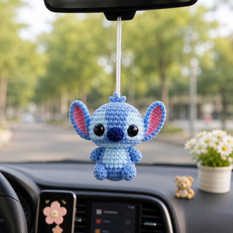 May include: A blue and light blue crochet Stitch car charm hanging from a car's rearview mirror. The charm has large ears with pink inner ears, black eyes, and a small nose. The charm is suspended by a white string.