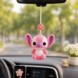 May include: A handmade crochet car charm featuring a pink and white character with large ears and a purple interior. The charm is suspended from a white string and includes a pink flower accent. The charm is hanging from a car's rearview mirror.