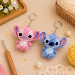 May include: Two handmade crochet keychains in the shape of a cartoon character, one pink and one blue. The pink keychain has purple accents, and the blue keychain has blue accents. Both have large black eyes and are attached to silver keyrings.