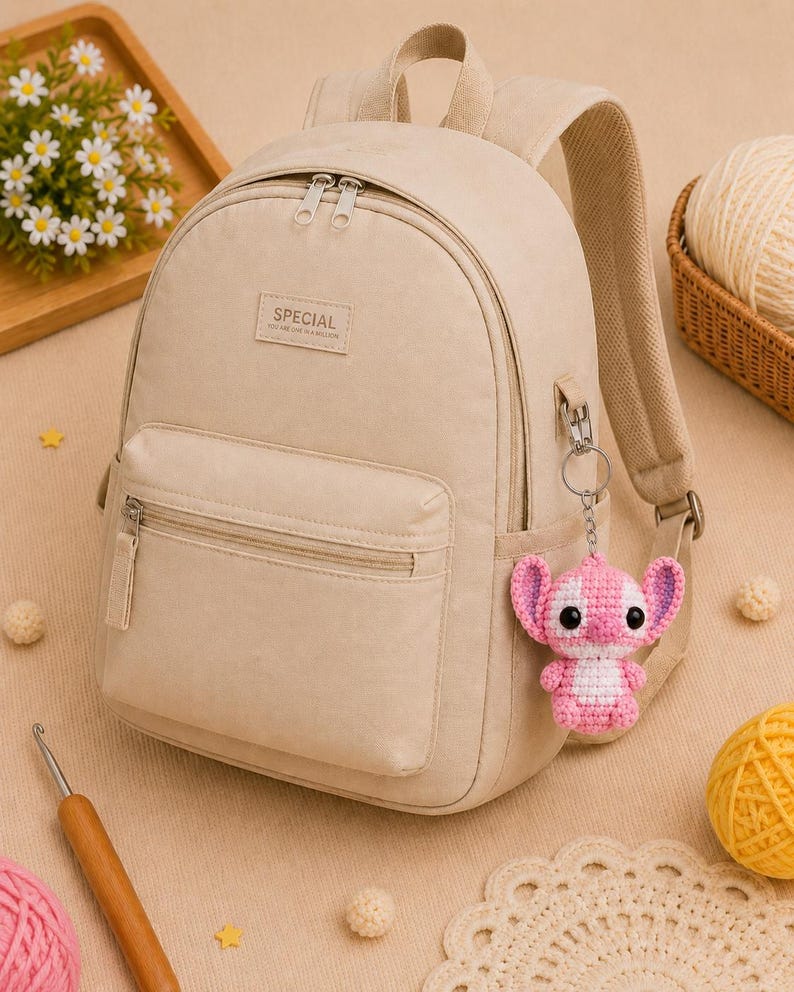 May include: Beige backpack with a front pocket and silver zippers. A small, pink and white crocheted Stitch keychain hangs from the side. The backpack has the word "SPECIAL" on the front.