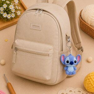 May include: Beige backpack with a front zippered pocket and the word "SPECIAL" on a rectangular patch. A blue and light blue crocheted Stitch keychain hangs from the side. The background includes yarn, crochet hook, and a tray of flowers.