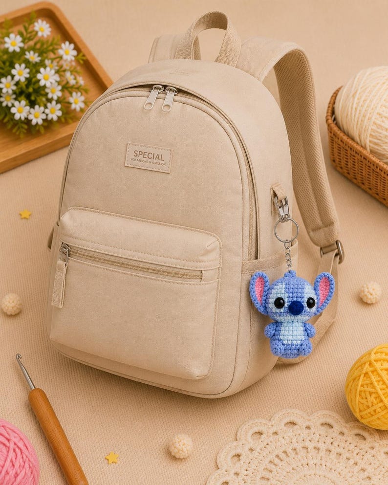 May include: Beige backpack with a front zippered pocket and the word "SPECIAL" on a rectangular patch. A blue and light blue crocheted Stitch keychain hangs from the side. The background includes yarn, crochet hook, and a tray of flowers.