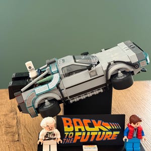Può includere: Un modello LEGO della macchina del tempo DeLorean di Ritorno al futuro, montato su un supporto nero con il logo del film e una targa California OUTATIME, accompagnato da due minifigure di Doc Brown e Marty McFly.