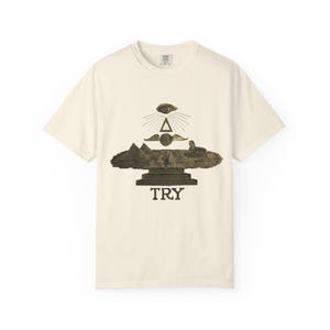 Könnte beinhalten: Cremefarbenes T-Shirt mit einem sepiafarbenen Aufdruck von Pyramiden, einer Sphinx und einem Auge über dem Wort "TRY". Das Design beinhaltet ein Auge, Flügel und ein Dreieck. Das Shirt hat einen Rundhalsausschnitt und kurze Ärmel.
