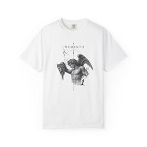 Könnte beinhalten: Ein weißes Kurzarm-T-Shirt mit einem schwarz-weißen Grafikdruck einer geflügelten Figur, die einen abgetrennten Kopf hält. Das Design umfasst geometrische Linien, einen Blitz und den Text MEMENTO MORI in einer Serifenschrift.