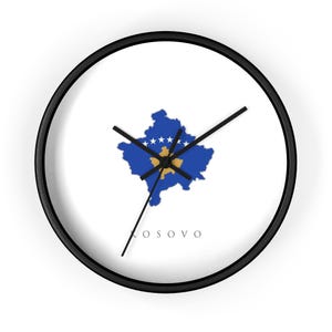 Könnte beinhalten: Eine runde Wanduhr mit schwarzem Rahmen und weißem Zifferblatt. In der Mitte befindet sich eine blaue Landkarte des Kosovo mit goldenem Umriss und sechs weißen Sternen in einem Bogen. Das Wort KOSOVO ist unten in schwarzen Großbuchstaben gedruckt.