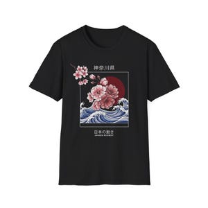 Könnte beinhalten: Ein schwarzes Kurzarm-T-Shirt mit einem Grafikdruck aus rosa Kirschblüten, blauen Meereswellen und einer roten Sonne. Das Design enthält den japanischen Text 神奈川県 und 日本の動き sowie den englischen Text JAPANESE MOVEMENT, eingerahmt in einem weißen Kasten.