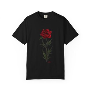 Könnte beinhalten: Schwarzes T-Shirt mit einem zentralen Motiv einer roten Rose und Cannabisblättern. Die Rose ist detailliert dargestellt, mit grünen Blättern und einem kleinen roten Tropfen am Stielansatz.