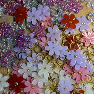 May include: A selection of flower-shaped keyrings in shades of red, pink, white, and purple. Some are solid, others glittery. Each keyring has a gold-coloured ring. The keyrings are scattered.