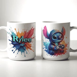 May include: Two white ceramic mugs. One mug features the name "Rylee" in teal letters against a colourful paint splash design. The other mug has a blue cartoon character wearing a hoodie and holding a game controller, with a paint splash background.
