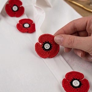 Può includere: Quattro decorazioni a forma di papavero lavorate all'uncinetto in rosso con centri neri e bottoni bianchi a quattro fori fissati al centro di ogni fiore. I papaveri sono disposti su una camicia bianca liscia con colletto.