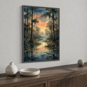 May include: A framed watercolor painting depicts a serene swamp scene. Tall trees with Spanish moss line the water's edge, reflecting in the calm water. The sky transitions from orange to blue, creating a tranquil atmosphere. The artwork is displayed on a wooden surface.