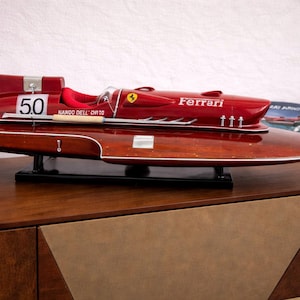 Może przedstawiać: Model łodzi wyścigowej w kolorze czerwonym i drewnianym, z logo Ferrari, napisem Nando dell'Orto oraz numerem 5.0 na boku. Łódź ma smukły, aerodynamiczny kształt ze spiczastym dziobem, czerwoną kabiną i czarną podstawką ekspozycyjną.