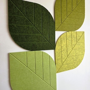 May include: Four leaf-shaped wall panels made of felted material in varying shades of green. Each panel features a central vein and parallel diagonal lines etched into the surface, creating a textured, layered arrangement of dark and light green.