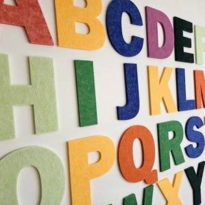 Könnte beinhalten: Eine Sammlung von sechsundzwanzig einzelnen Filzbuchstaben, die das Alphabet von A bis Z darstellen. Jeder Buchstabe hat eine eigene Volltonfarbe, darunter Rot, Gelb, Blau, Lila, Schwarz, Hellgrün, Dunkelgrün, Orange und Pink.
