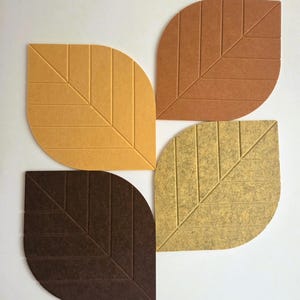May include: Four leaf-shaped wall panels made of felted material, each featuring a central vein and diagonal etched lines. The set includes one yellow, one light brown, one dark brown, and one mottled beige leaf, all arranged in a cluster.
