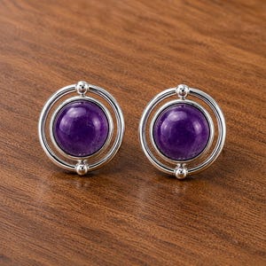 May include: A pair of stud earrings featuring a smooth, round, deep purple amethyst cabochon stone set within a polished silver-toned circular frame. Each earring has a small silver sphere at the top and bottom of the outer ring structure.