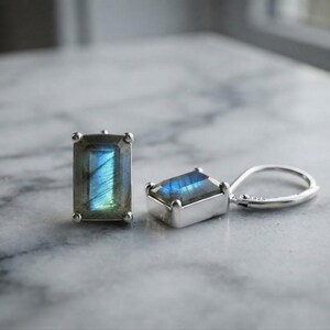 May include: A pair of rectangular labradorite earrings set in sterling silver. Each earring features a faceted labradorite stone displaying a blue iridescent flash, held by four prongs in a silver setting with a secure lever back closure.