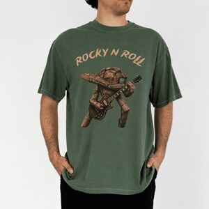 May include: Green t-shirt with the text "Rocky N Roll" above a brown, guitar-playing creature. The creature has a rock-like head and body. The shirt is a solid, muted green color.