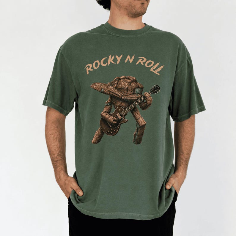 May include: Green t-shirt with the text "Rocky N Roll" above a brown, guitar-playing creature. The creature has a rock-like head and body. The shirt is a solid, muted green color.
