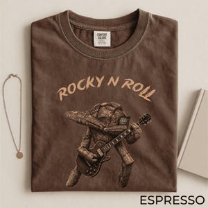 May include: Espresso-colored t-shirt with the words "ROCKY N ROLL" in a light brown font. The shirt features a graphic of a rock-like creature playing a guitar. The shirt has a crew neck and a tag that says "Comfort Colors".