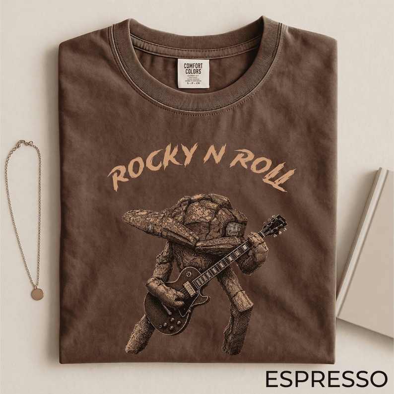 May include: Espresso-colored t-shirt with the words "ROCKY N ROLL" in a light brown font. The shirt features a graphic of a rock-like creature playing a guitar. The shirt has a crew neck and a tag that says "Comfort Colors".