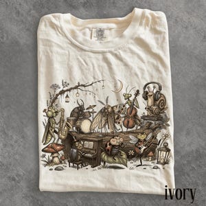 Może przedstawiać: T-shirt w kolorze kości słoniowej z grafiką przedstawiającą owady grające na instrumentach muzycznych, w tym konika polnego na saksofonie, żuka na perkusji, ćmę na wiolonczeli, ślimaka w słuchawkach, biedronkę na pianinie i pszczołę na gitarze.