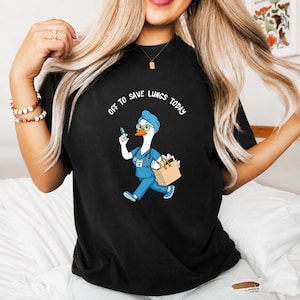 May include: Black t-shirt featuring a cartoon goose dressed as a nurse, holding a syringe and a tote bag. The goose wears a blue uniform, glasses, and a nurse's cap. Text on the shirt reads "OFF TO SAVE LUNGS TODAY."