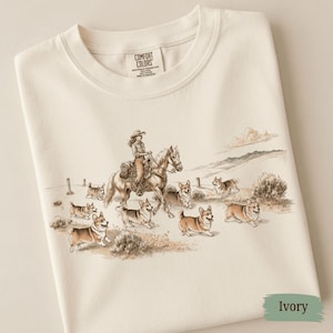 May include: An ivory t-shirt featuring a graphic print of a person riding a horse through a landscape with several Corgi dogs running alongside. The shirt has a Comfort Colors brand label at the inner neckline and a short-sleeved crew neck design.
