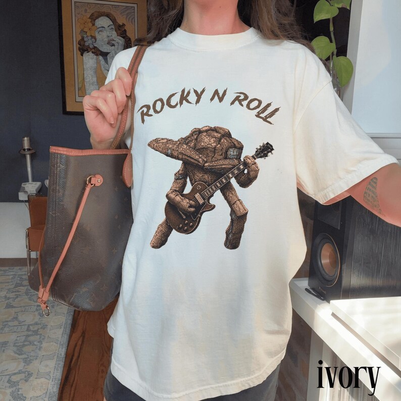 May include: Ivory-colored t-shirt featuring a brown graphic of a rock-like creature playing a guitar, with the text "ROCKY N ROLL" above the graphic. The shirt is a relaxed fit.
