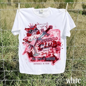 May include: A white t-shirt featuring a graphic print of a cat wearing goggles and a crown, a sword, a martini glass, and red watercolor splashes. The text reads: Mama needs a night off, DESPERADO CLUB, home of the Dirty Shirly, SO FUN IT HURTS, ENTRANCE IN REAR.