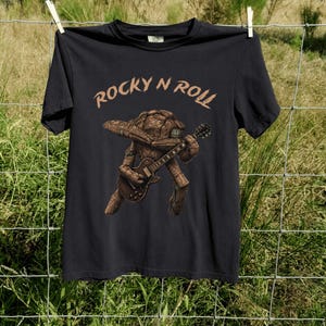 May include: Black t-shirt with the text "ROCKY N ROLL" in a brown font. The shirt features a graphic of a rock-like creature playing a brown electric guitar. The shirt is hanging on a clothesline.