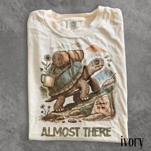 Peut inclure: Un t-shirt ivoire avec une illustration de tortue portant un sac de couchage roulé et une tasse sur son dos. La tortue marche sur un sentier rocheux vers des montagnes. Un panneau indique Summit: 14 mi. Le texte ALMOST THERE est imprimé en bas.