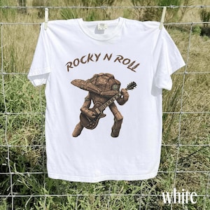 May include: White t-shirt with a brown illustration of a rock-and-roll creature playing a guitar. The text "ROCKY N ROLL" is above the illustration. The word "white" is at the bottom right of the shirt.
