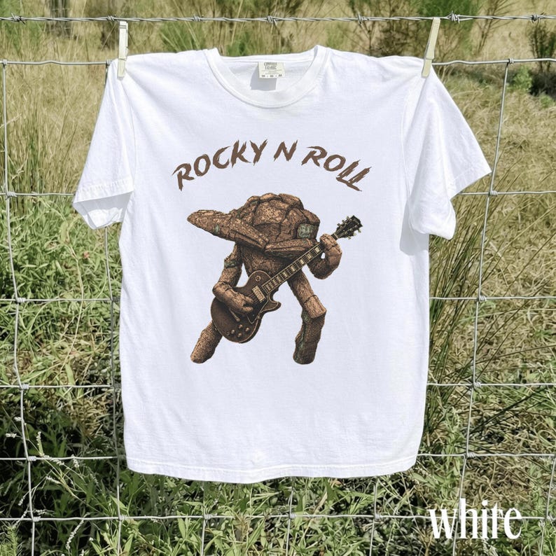 May include: White t-shirt with a brown illustration of a rock-and-roll creature playing a guitar. The text "ROCKY N ROLL" is above the illustration. The word "white" is at the bottom right of the shirt.