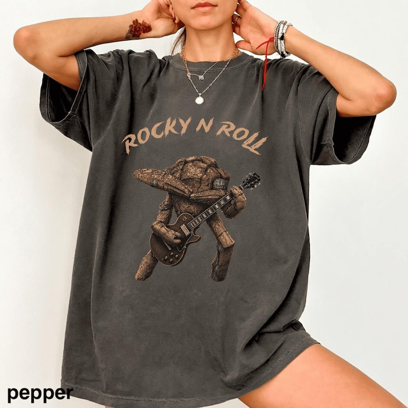 May include: Dark grey t-shirt with the text "ROCKY N ROLL" in a brown font. The shirt features a brown illustration of a rock-like creature playing a guitar. The word "pepper" is printed at the bottom left.