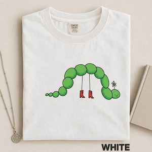 Op de afbeelding: Een wit t-shirt met een afbeelding van een groene gesegmenteerde rups met een klein bloemetje op zijn kop. De rups heeft twee dunne zwarte benen met rode laarzen aan. Het shirt heeft een Comfort Colors merklabel bij de binnenhalslijn.