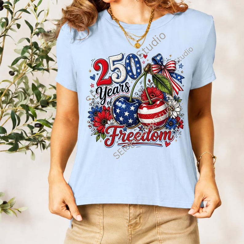 May include: A light blue short-sleeved t-shirt featuring a graphic print of two cherries with American flag patterns, a red, white, and blue bow, and the text 250 Years of Freedom surrounded by red and blue flowers and small stars.